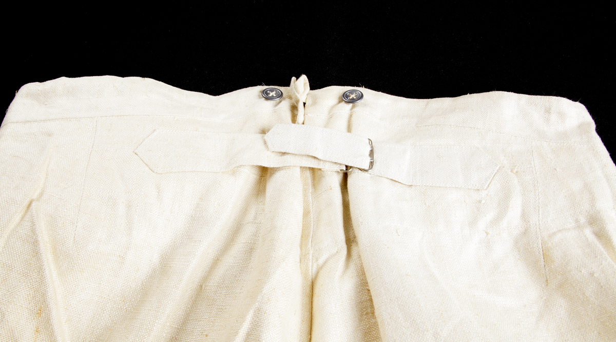 Uniformsbyxor (sommaruniform). Rak modell med pressveck, i ljusbeige, grov bomull. Knäppgylf med tre blyknappar. Två sidofickor, sju blyknappar i midjan (bärs med hängslen), sprund baktill i linningen där byxornas vidd kan regleras med ett spänne strax nedanför.