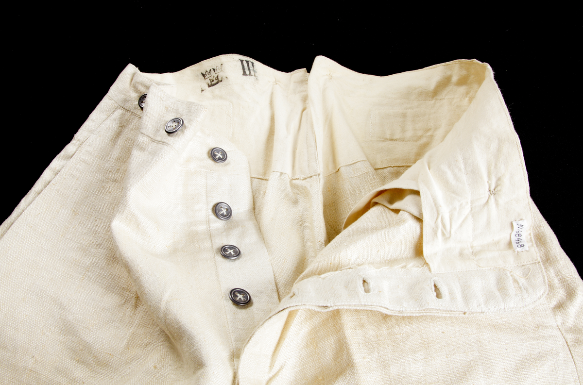 Uniformsbyxor (sommaruniform). Rak modell med pressveck, i ljusbeige, grov bomull. Knäppgylf med tre blyknappar. Två sidofickor, sju blyknappar i midjan (bärs med hängslen), sprund baktill i linningen där byxornas vidd kan regleras med ett spänne strax nedanför.