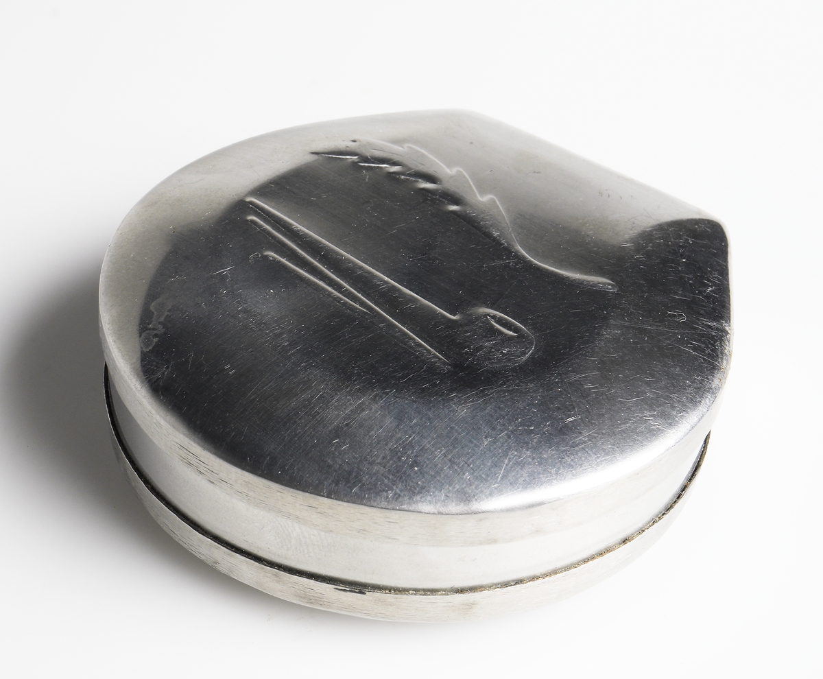 Dosa i metall. Lock med gångjärn. På locket ett pressat motiv föreställande en pipa och ett tobaksblad.