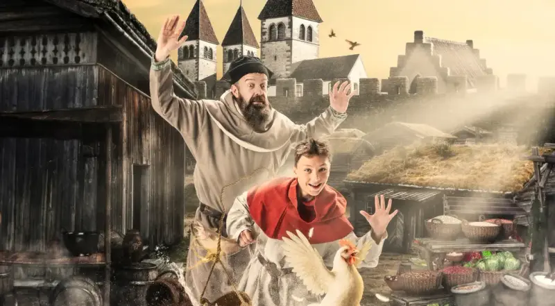 Mann og gutt i middelalderklær som løper etter hverandre. Middelalderby og kirke i bakgrunnen flaksende høne foran