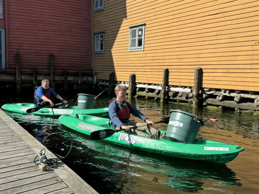 To menn sitter i hver sin grønne kajakk på vei ut fra museet for å samle søppel som flyter