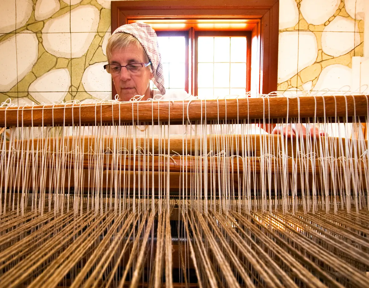 En äldre kvinna med huckle sitter och väver i en vävstol.