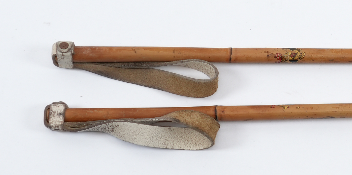 Bambusstavar med skinnreimer som handtak. Skinnreimene er kvite og festa til bambusstavane med ei nagle. På bambusstavane er det spor etter klistermerker med ukjend motiv, men med påskrift "Standard" og "Made in Norway". Trinser av bambus og lær. 