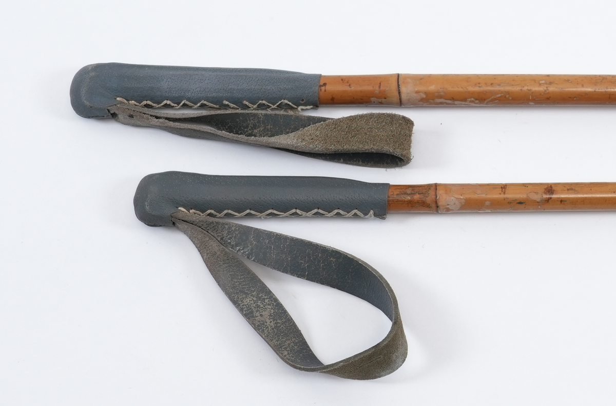 Eit par eldre skistavar i tre, truleg bambus, med påsette handtak i gråblått lær. Handtaka er sydd langs sidene med lys snøring og har fastmonterte handstropper i lær, festa med nagle gjennom ein opning i handtaket. Trisene nedst på skistavane har bambusring festa til skistaven med fire lærreimer.