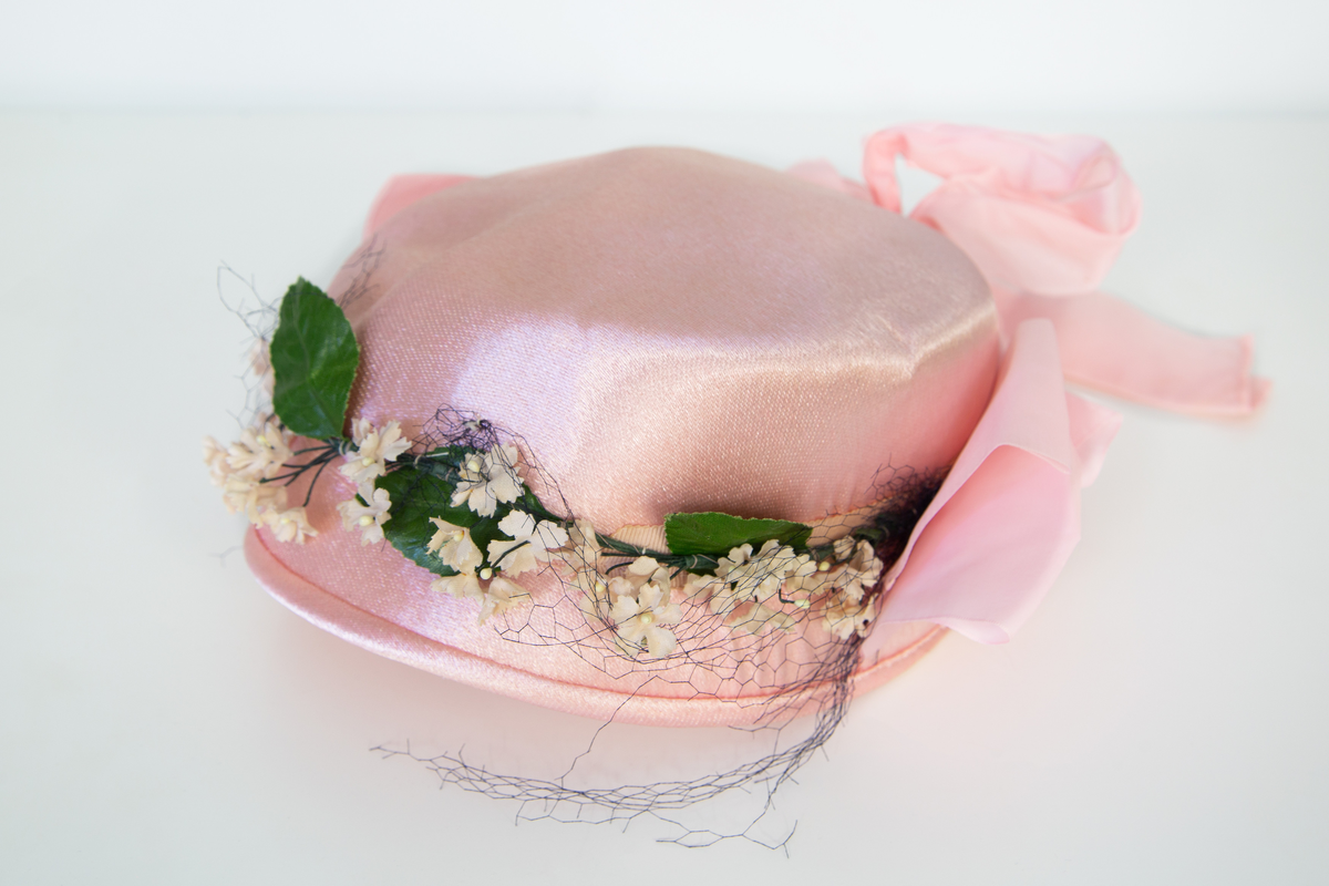 Rosa damhatt dekorerad med en ranka av vita tygblommor samt gröna blad, svart tyll och två rosa band fästa strax bakom öronen på vardera sidan. Till Hatten hör ett par vita  näthandskar försedda med en liten vit knapp. 