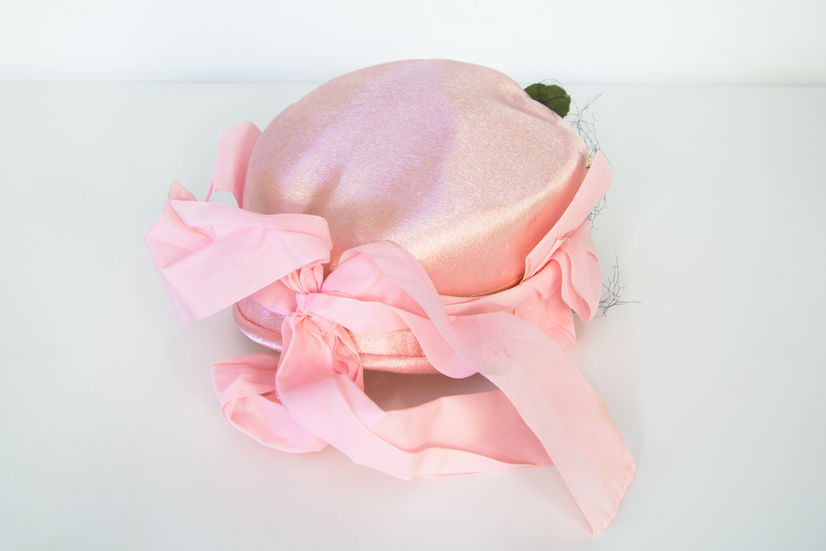 Rosa damhatt dekorerad med en ranka av vita tygblommor samt gröna blad, svart tyll och två rosa band fästa strax bakom öronen på vardera sidan. Till Hatten hör ett par vita  näthandskar försedda med en liten vit knapp. 