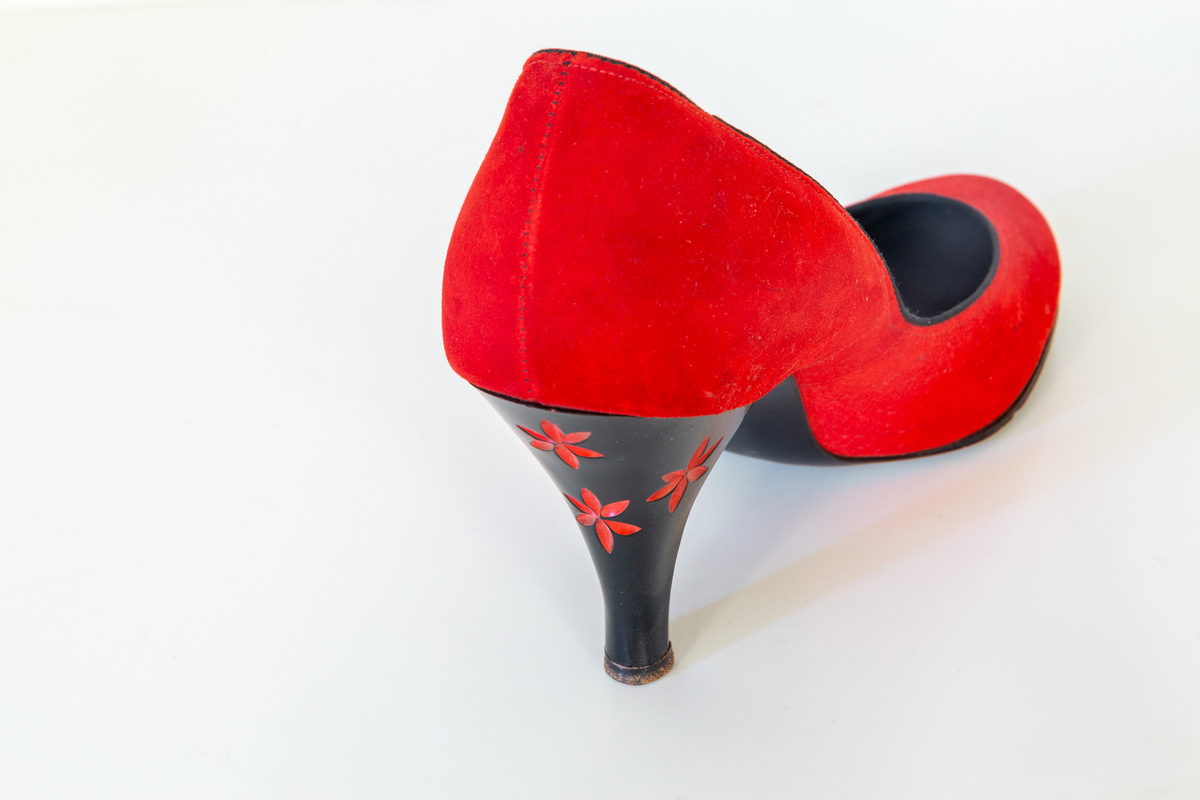 Högklackade skor sk pumps i röd mocka. De svarta klackarna är dekorerade med fyra, ur klacken utskurna röda blommor med vardera sex kronblad. Storlek 36.