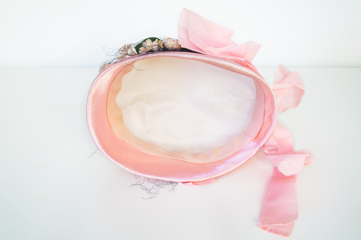 Rosa damhatt dekorerad med en ranka av vita tygblommor samt gröna blad, svart tyll och två rosa band fästa strax bakom öronen på vardera sidan. Till Hatten hör ett par vita  näthandskar försedda med en liten vit knapp. 