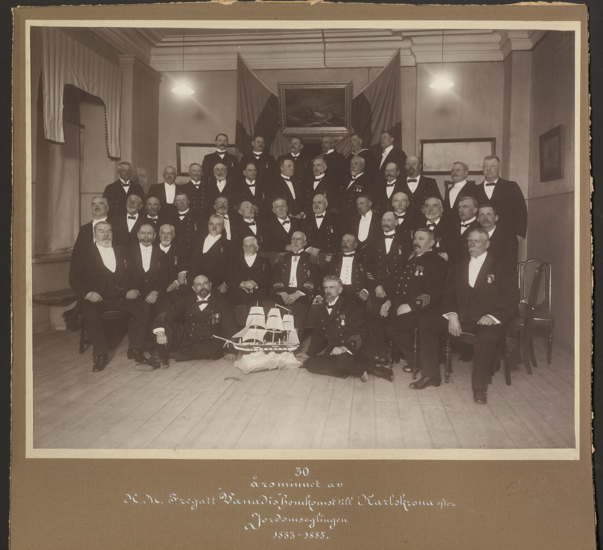 Bilden föreställer ett gruppfoto under 30 årsminnet av H.M. Fregatt Vanadis hemkomst till Karlskrona efter jordomseglingen 1883-1885.
Personen stående till vänster vid gardinens högra mitt är flaggskeppare Ernst Fhal. I mitten amiral Lagercrantz. Sittande till vänster om båten flaggskeppare Mortensson. Sittandes längst fram till vänster i civila kläder är mest troligt Frans Wilhelm Elftman.