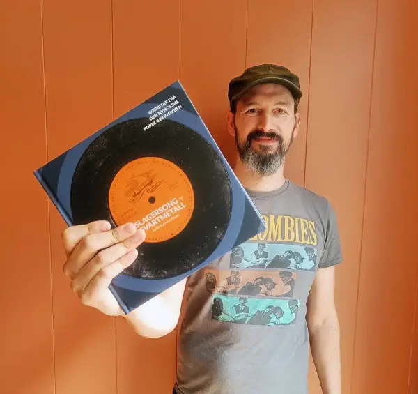 Arild Torvund Olsen med skuggelue og med ei bok i handa framom ein oransje vegg.