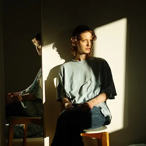 Anna With sitjande på ein krakk framfor ein spegel, i delvis skugge med lys frå vindauge