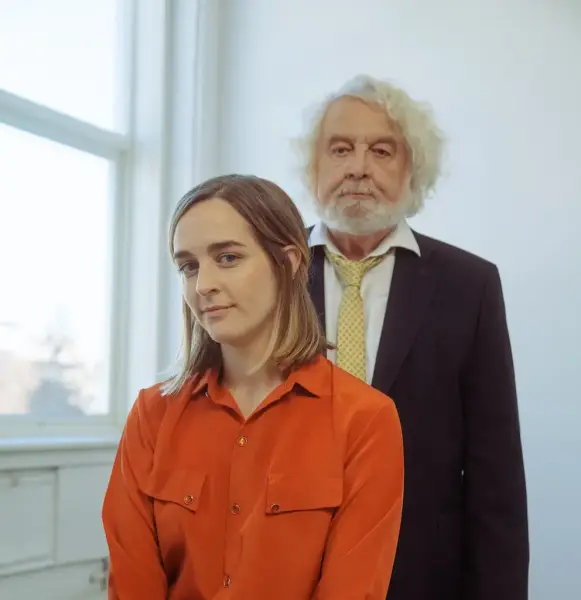 Ine Hoem med raud skjorte og Edvard Hoem med dress og gult slips, framom eit vindauge i eit kvitt rom