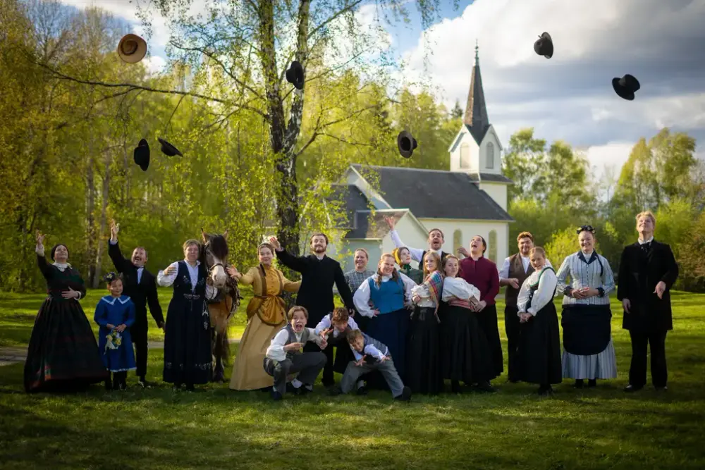 Alle skuespillerne i kostymer poserer foran utvandrerkirken og kaster hattene i luften