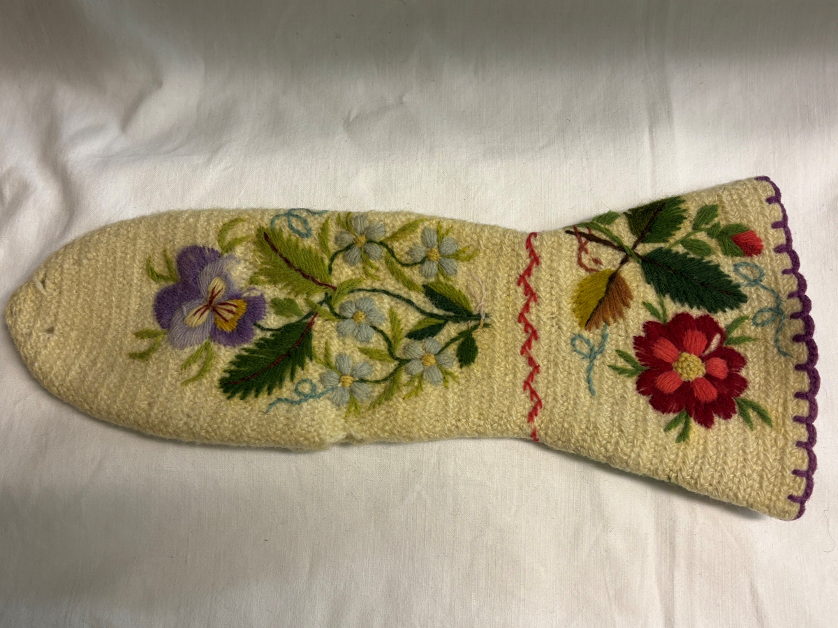 Vit nålbunden vante med broderi i blomstermotiv. Lång mudd med dekorationer runt om. Även dekorationer på tummarna. Högervante.