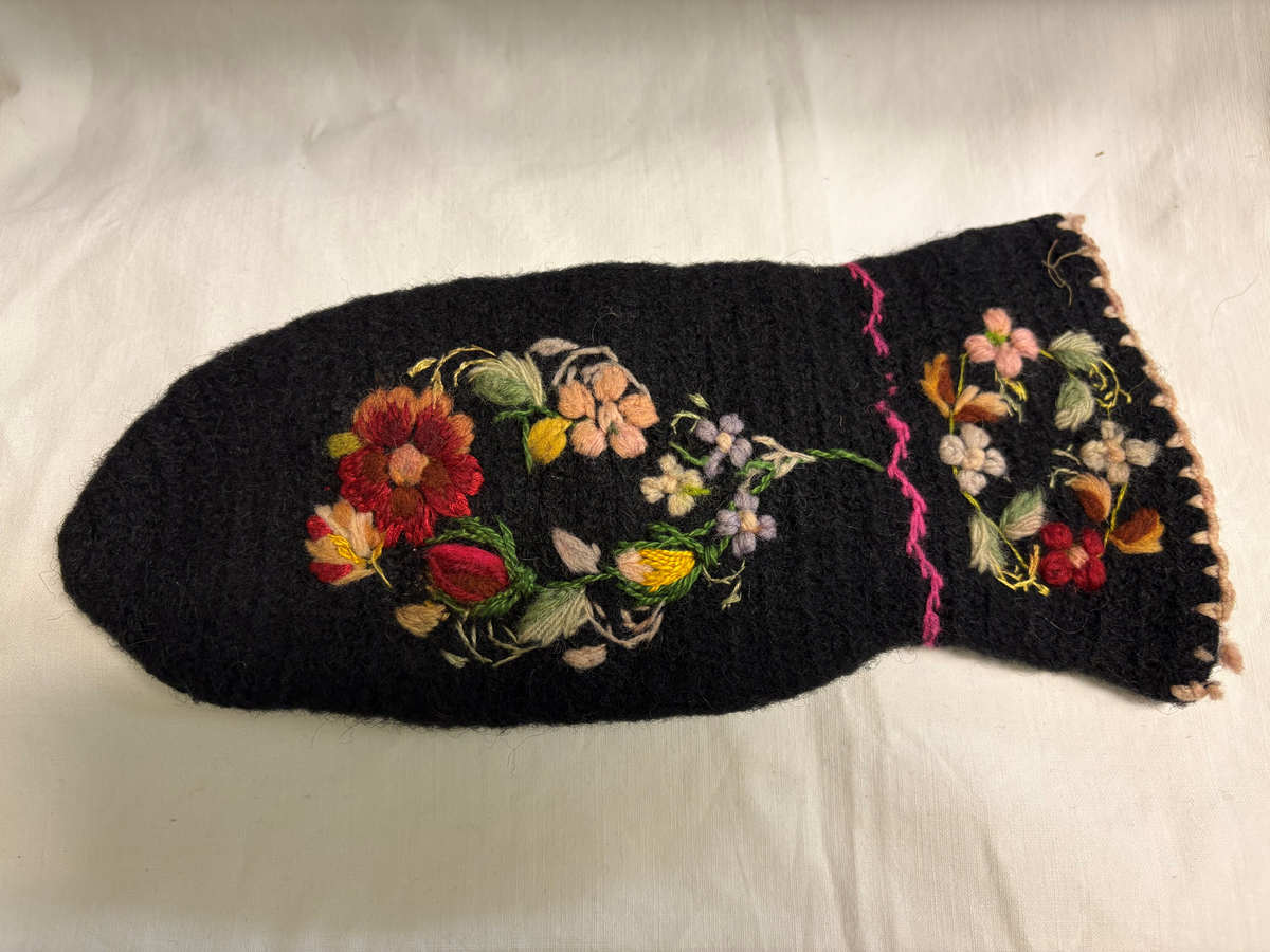 Svart nålbunden vante med broderi i blomstermotiv. Lång mudd med dekorationer runt om. Även dekorationer på tummarna. Vänstervante.