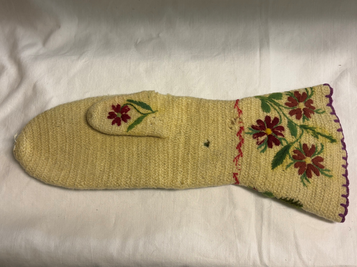 Vit nålbunden vante med broderi i blomstermotiv. Lång mudd med dekorationer runt om. Även dekorationer på tummarna. Högervante.