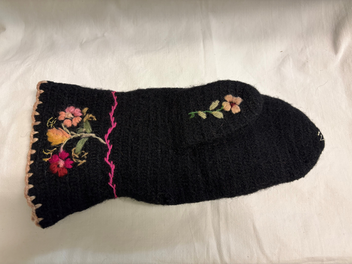 Svart nålbunden vante med broderi i blomstermotiv. Lång mudd med dekorationer runt om. Även dekorationer på tummarna. Vänstervante.