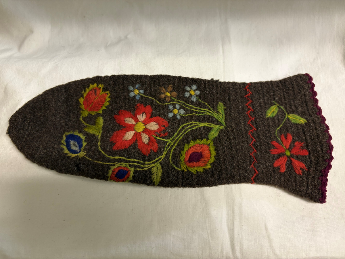 Grå nålbunden vante med broderi i blomstermotiv. Lång mudd med dekorationer runt om. Även dekorationer på tummarna. Högervante.
