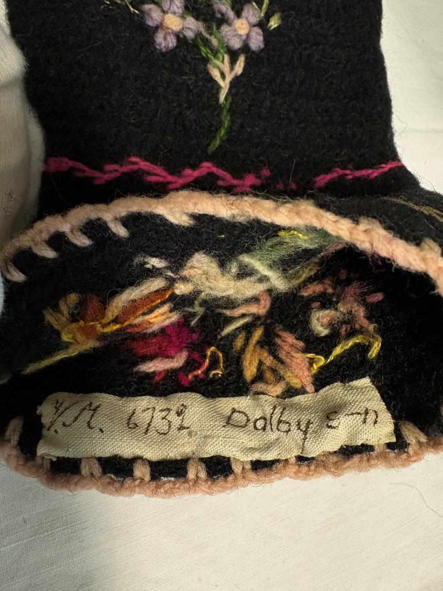 Svart nålbunden vante med broderi i blomstermotiv. Lång mudd med dekorationer runt om. Även dekorationer på tummarna. Vänstervante.