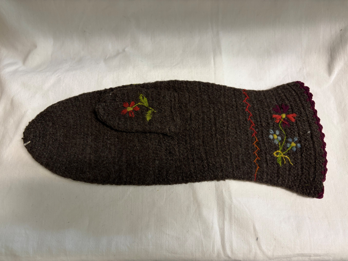 Grå nålbunden vante med broderi i blomstermotiv. Lång mudd med dekorationer runt om. Även dekorationer på tummarna. Högervante.