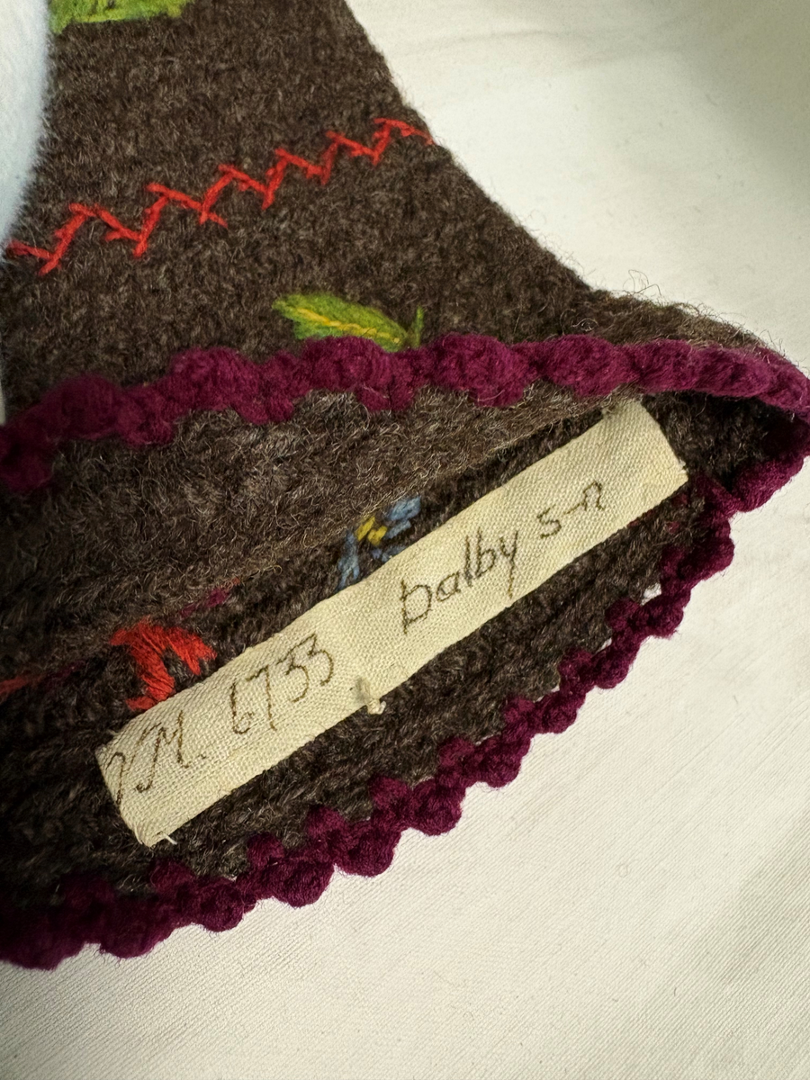 Grå nålbunden vante med broderi i blomstermotiv. Lång mudd med dekorationer runt om. Även dekorationer på tummarna. Högervante.