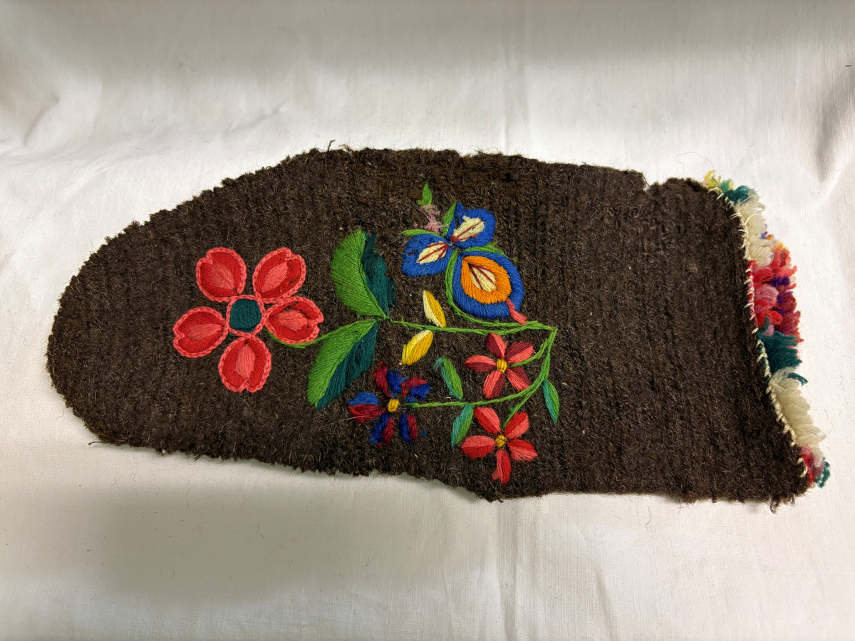 Tumvante av brunt ullgarn med broderier i schattersöm och kedjesöm i färgerna: rött, vitt, blått, rosa, gult och grönt. Flossad kant i samma färger.