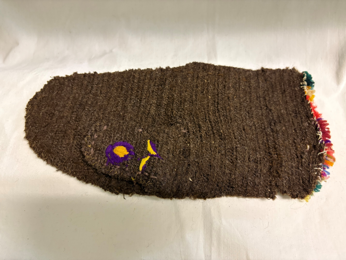 Tumvante av brunt ullgarn med broderier i schattersöm och kedjesöm i färgerna: rött, vitt, blått, rosa, gult och grönt. Flossad kant i samma färger.