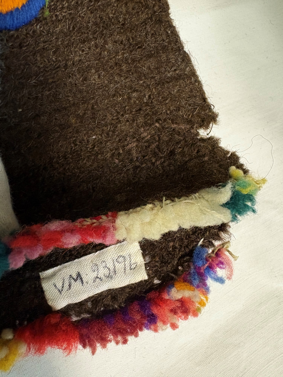 Tumvante av brunt ullgarn med broderier i schattersöm och kedjesöm i färgerna: rött, vitt, blått, rosa, gult och grönt. Flossad kant i samma färger.