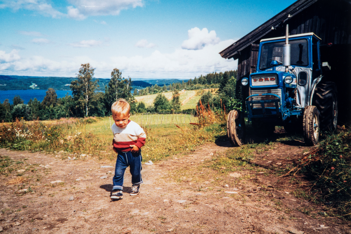 Stange, Espa, Kjeldsrud gård. Sommer 1985. Øyvind Romsaas Borgersen 2 år. Til høyre skimtes den gamle smia bak traktoren. Utsikt til Tangenvika i bakgrunnen. Ford traktor.
