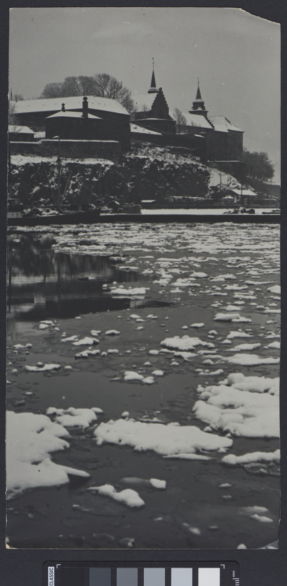 Utsikt til en festning (Akershus festning?) ved vannkanten. Bakken, vegetasjonen rundt og alle flater er dekket med snø, og på vannet flyter det isflak.