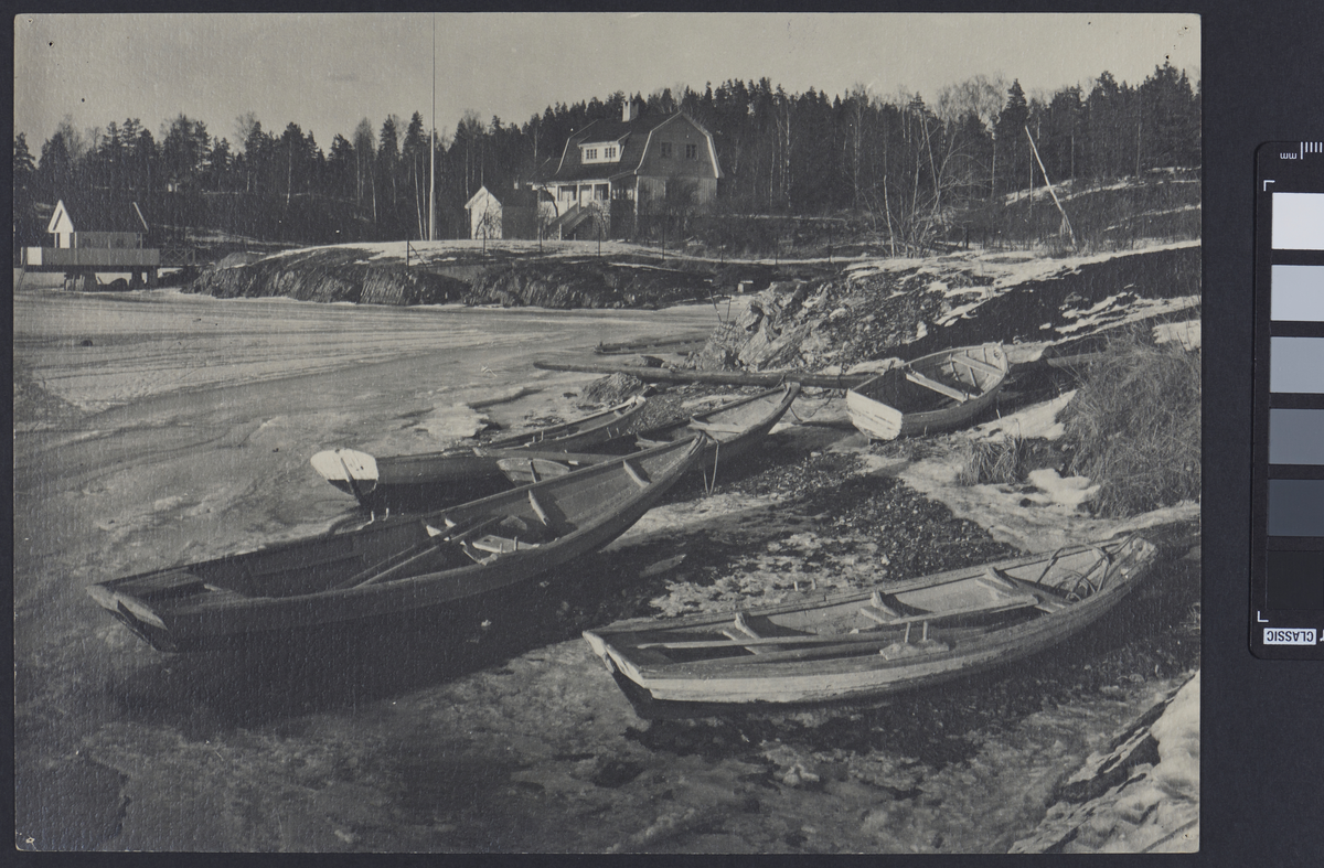 En strand hvor flere trebåter ligger på land for vinteren. Vannet er isete og bakken er delvis dekket av et lag snø. I bakgrunnen er det et to etasjes hus og et mindre hus med veranda som står ute i vannet. En skog strekker seg bortover bak husene.