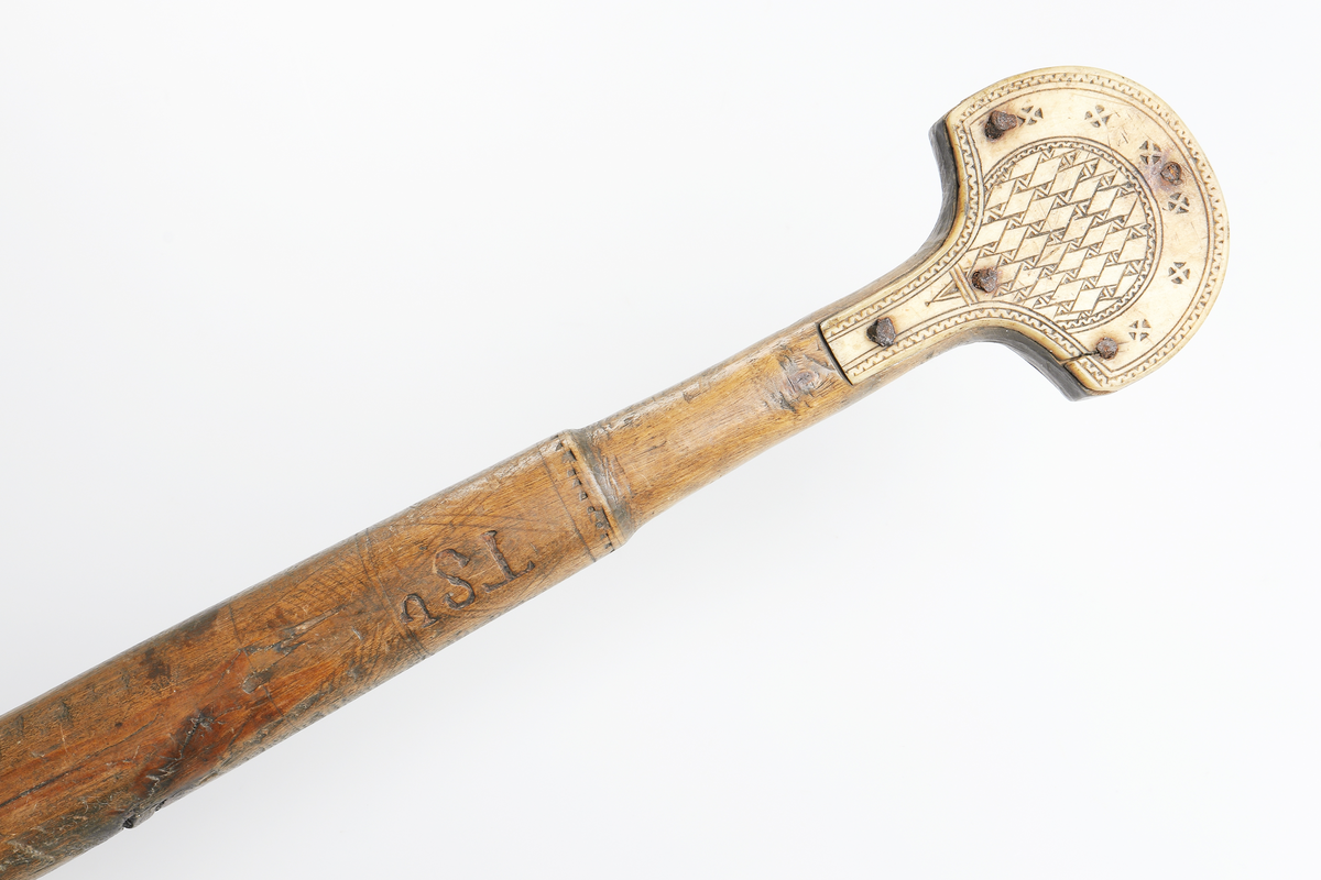 Käpp av trä med benförsedd knopp. Benstyckerna, ett på vardera sidan, är dekorerade med geometriska figurer, fastsatta med fem spikar. Inuti käppen har man lagt i stenar eller liknande så att den skramlar när man rör den. Från Gällivare.