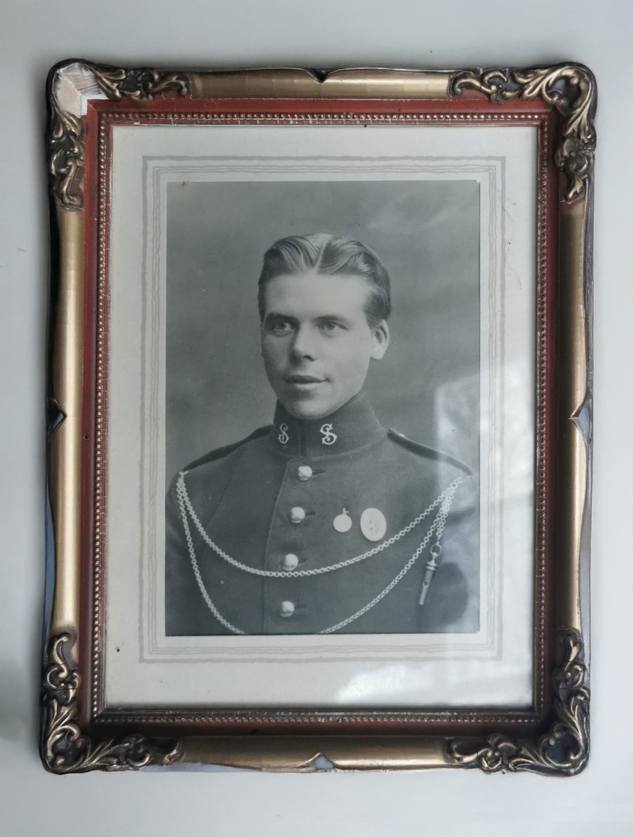 Fotografi i ramme i høydeformat. Brystbilde, 3/4 profil, dreiing venstre. Militær iniform. Emblemet på kraven: Gardens musikkorps. Dobbel lenke over brystet som ender i en fløyte. To medaljer på brystet, den ene med riksvåpenet.
Treramme, brunmalt med stukkornamenter i hjørnene, og perlestav inn mot glasset. Lite stykke av stukkornamentet falt av i venstre, øvre hjørne. Baksiden: Papp med bordstøtte. 
På bordstøtten er påklistret rød lapp med gullbokstaver.
Øverst bak, hempe for opphenging.