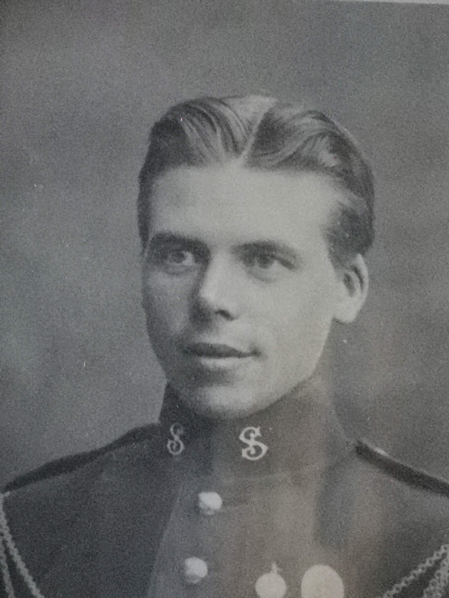 Fotografi i ramme i høydeformat. Brystbilde, 3/4 profil, dreiing venstre. Militær iniform. Emblemet på kraven: Gardens musikkorps. Dobbel lenke over brystet som ender i en fløyte. To medaljer på brystet, den ene med riksvåpenet.
Treramme, brunmalt med stukkornamenter i hjørnene, og perlestav inn mot glasset. Lite stykke av stukkornamentet falt av i venstre, øvre hjørne. Baksiden: Papp med bordstøtte. 
På bordstøtten er påklistret rød lapp med gullbokstaver.
Øverst bak, hempe for opphenging.