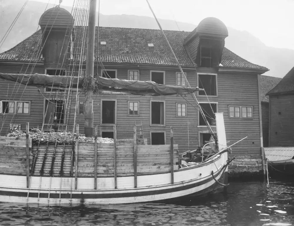 Sorthvitt bilde viser en fullastet nordlandsbåt foran en sjøbod