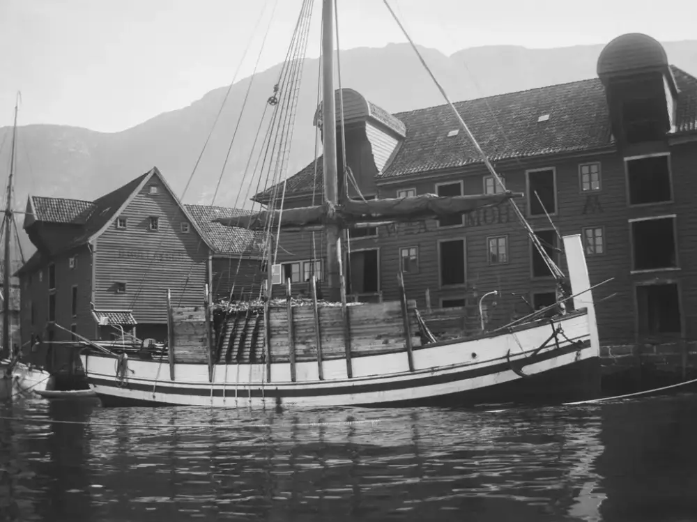 Bildet i sort-hvitt viser en nordlandsbåt lastet med tørrfisk foran sjøboder