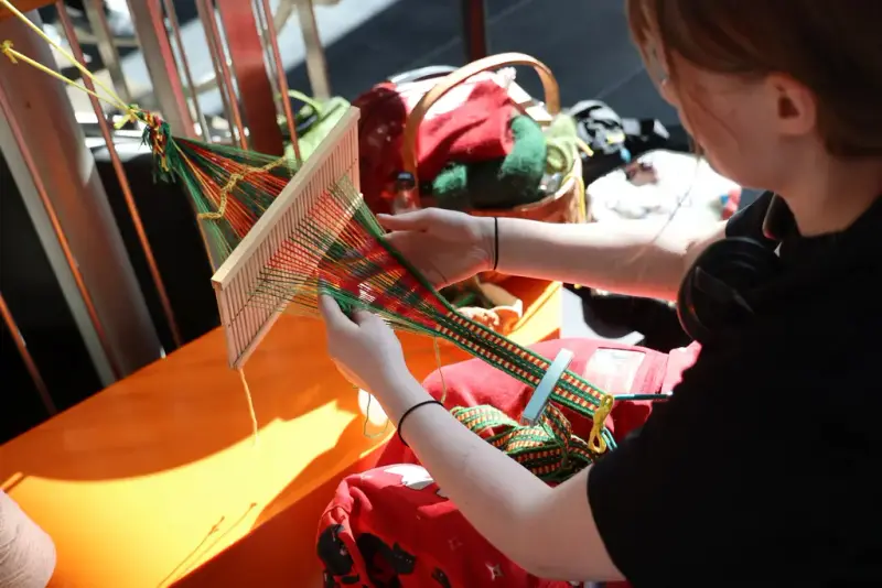 En person vever et fargerikt bånd med grindvev på et bord etter skogfinsk tradisjon. Garn og ferdige bånd ligger i en kurv i baggrunden.