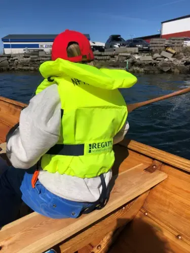 Vi ser et barn med stor gul redningsvest bakfra, mens barnet ror i en tradisjons robåt