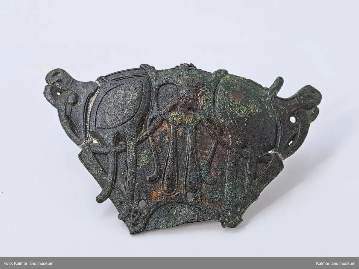 KLM 11464. Spänne(?). Gjuten välvd del av brons. Ornering med långsträckta detaljer där vissa delar av orneringen är genombruten och fyra hål ser ut att ha varit del av någon typ av fästanordning. På baksidan en anhopning av korrosion med del av vad som troligen är en länk av en kedja. Länken har oval form och är cirka 1,3 cm lång. I samma anhopning sitter en mindre bit, eventuellt en benbit. Spännet saknar delar samt har äldre lagningar på båda sidor, synliga på baksidan.