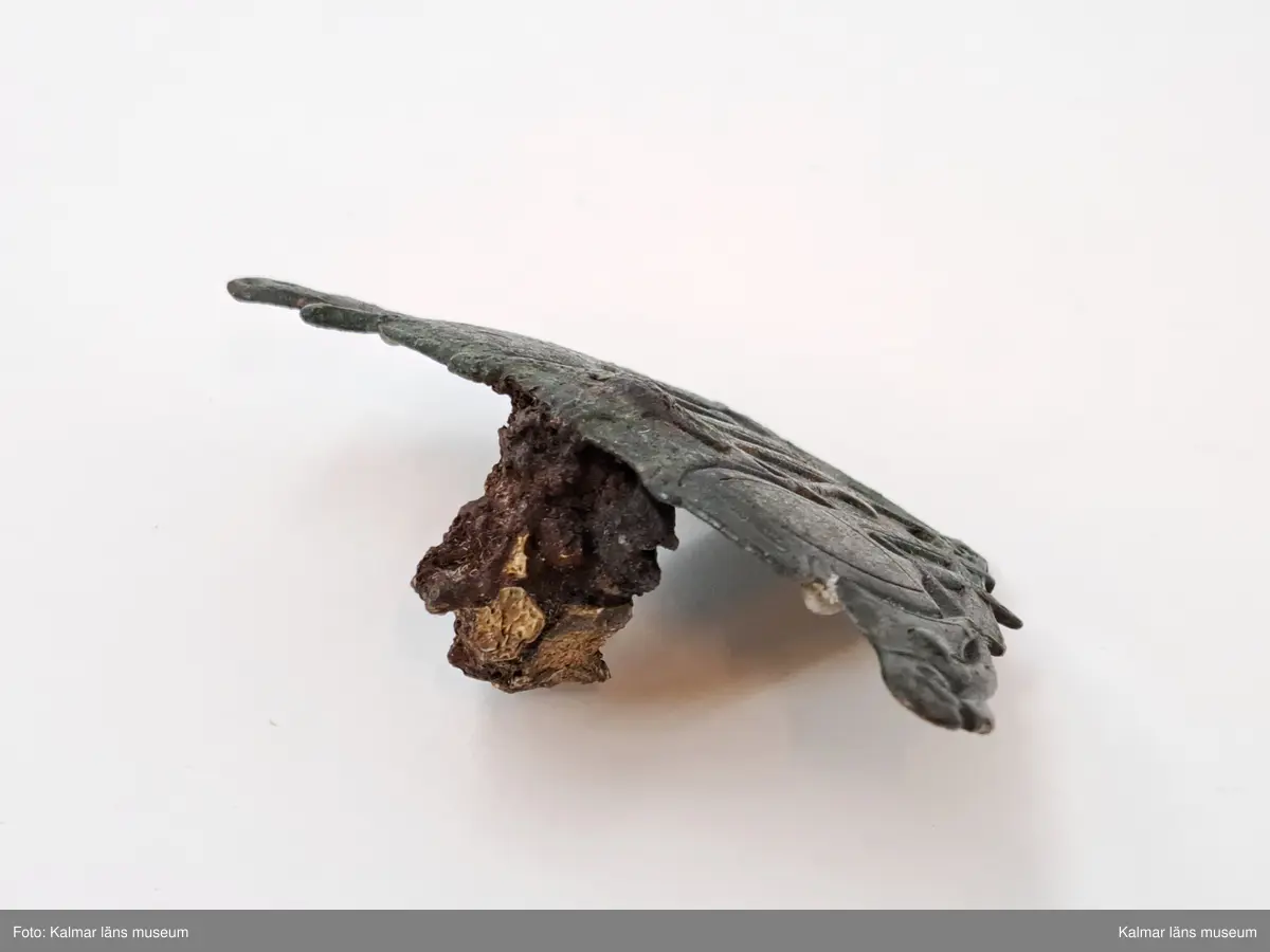 KLM 11464. Spänne(?). Gjuten välvd del av brons. Ornering med långsträckta detaljer där vissa delar av orneringen är genombruten och fyra hål ser ut att ha varit del av någon typ av fästanordning. På baksidan en anhopning av korrosion med del av vad som troligen är en länk av en kedja. Länken har oval form och är cirka 1,3 cm lång. I samma anhopning sitter en mindre bit, eventuellt en benbit. Spännet saknar delar samt har äldre lagningar på båda sidor, synliga på baksidan.