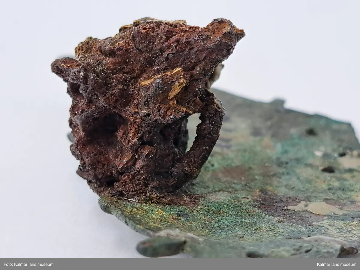 KLM 11464. Spänne(?). Gjuten välvd del av brons. Ornering med långsträckta detaljer där vissa delar av orneringen är genombruten och fyra hål ser ut att ha varit del av någon typ av fästanordning. På baksidan en anhopning av korrosion med del av vad som troligen är en länk av en kedja. Länken har oval form och är cirka 1,3 cm lång. I samma anhopning sitter en mindre bit, eventuellt en benbit. Spännet saknar delar samt har äldre lagningar på båda sidor, synliga på baksidan.