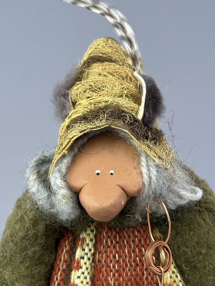 Krokig trollgumma med stora fötter och händer.  Är klädd i randig vävd kjol och grön jacka. Har lavbeklädd mössa med fjäder, lavbeklädda skor samt örhänge av koppar.

