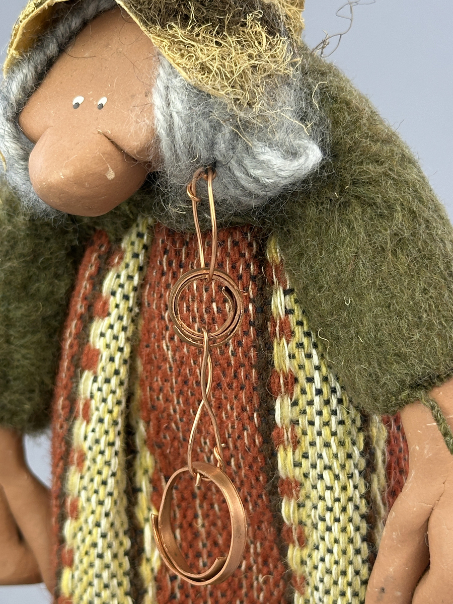 Krokig trollgumma med stora fötter och händer.  Är klädd i randig vävd kjol och grön jacka. Har lavbeklädd mössa med fjäder, lavbeklädda skor samt örhänge av koppar.

