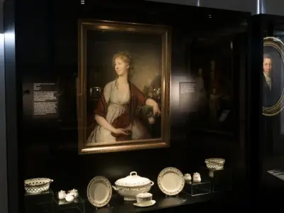 Portrett av en kvinne malt av Jens Juel, kledd i hvit kjole med rødbrun sjal, og holder en blomsterbukett i hånden. Utstilt i en monter sammen med hvite porselensgjenstander.