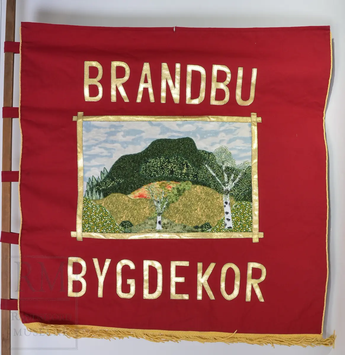 Rød bomullsfane med quiltet motiv
a: fane
b: stangpryd (harpe i messing)
c: bardunsnor m dusker
d: styresnor med håndgrep
e-g: stang og oppheng (e) i alu og messing
h: hvit lærsele
i: bæreveske/ oppbevaringspose i grått kunstskinn