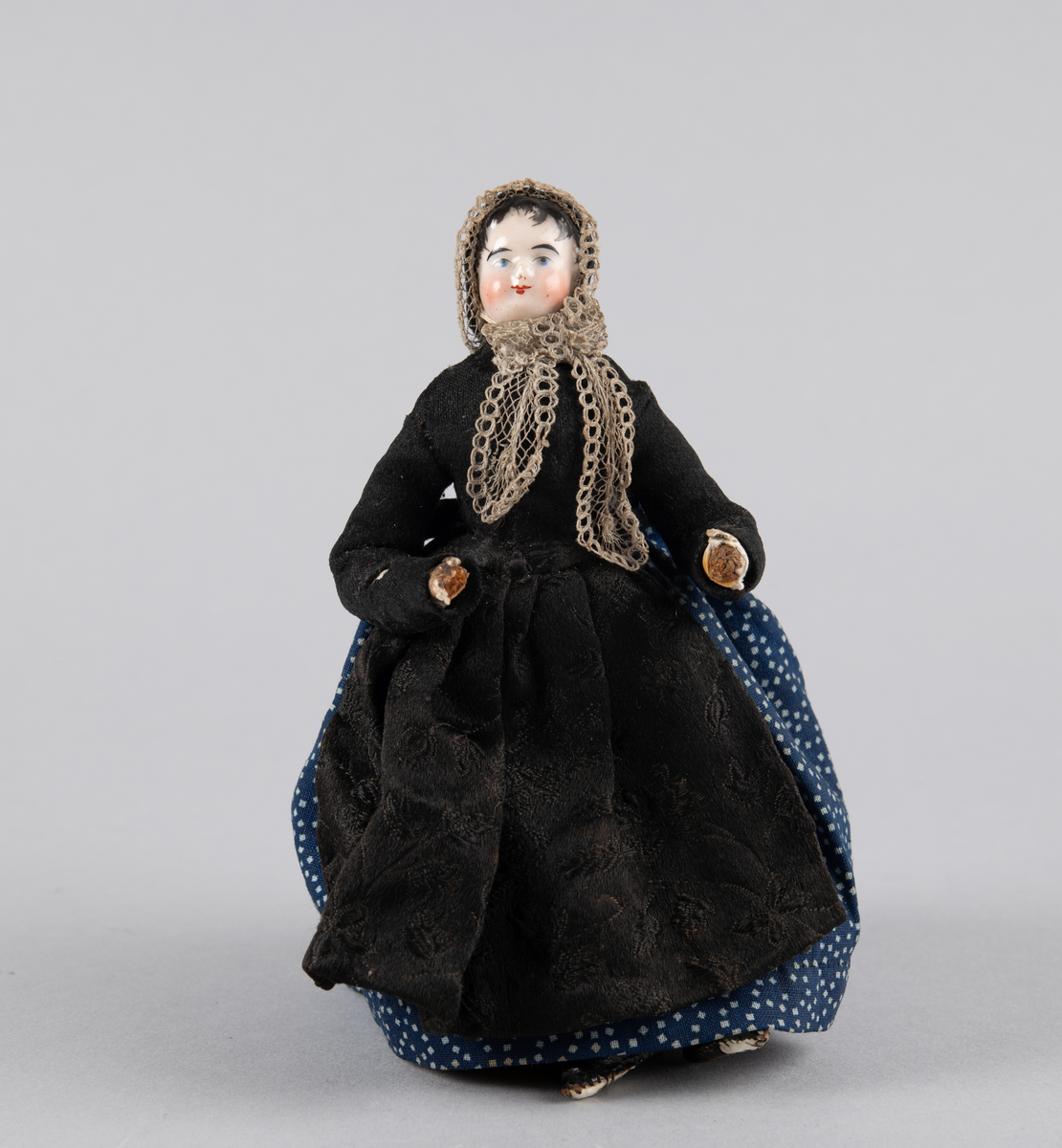 Liten dukke med porselenshode. Dame med prikkete skjørt og brokadeforkle. Hette med blonder foran og fløyelssløyfe bak. 2 underskjørt (ull og bomull)