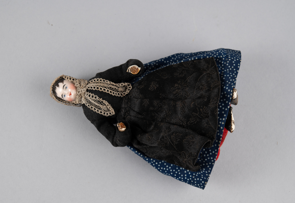Liten dukke med porselenshode. Dame med prikkete skjørt og brokadeforkle. Hette med blonder foran og fløyelssløyfe bak. 2 underskjørt (ull og bomull)