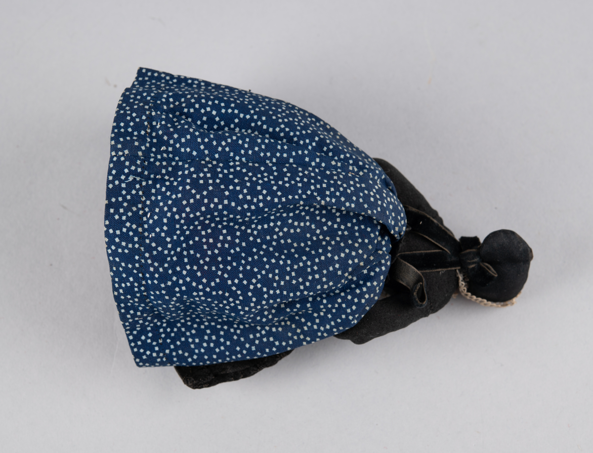 Liten dukke med porselenshode. Dame med prikkete skjørt og brokadeforkle. Hette med blonder foran og fløyelssløyfe bak. 2 underskjørt (ull og bomull)