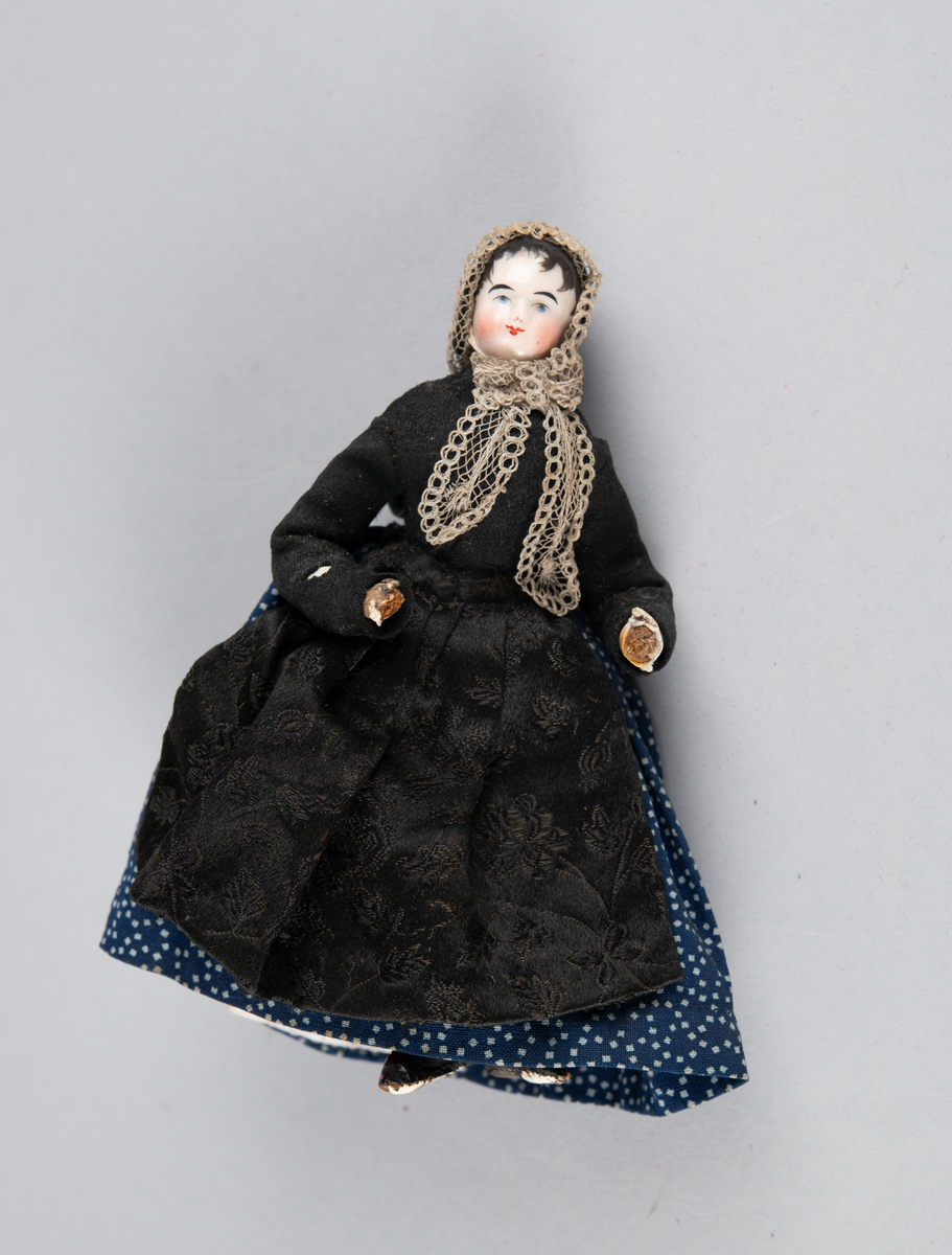 Liten dukke med porselenshode. Dame med prikkete skjørt og brokadeforkle. Hette med blonder foran og fløyelssløyfe bak. 2 underskjørt (ull og bomull)