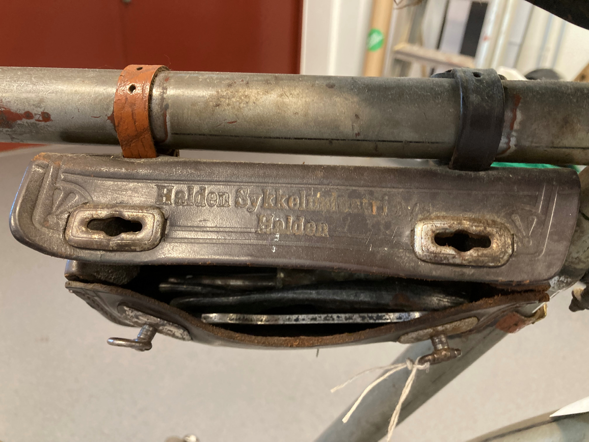 Herresykkel i metall, med sete av lær, lykt og bjelle. Påmontert verktøyeske i lær ved sete. Festet ved bagasjebrett 2 rødmalte halvsirkelformede kasser i tre, med en brannslange i hver.